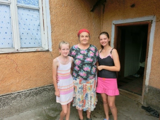 Drie mensen staan buiten voor een oranjebruine muur; een oudere vrouw in een jurk tussen twee meisjes. Raam en deur achter hen.