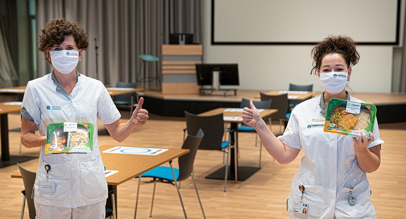 Twee zorgmedewerkers in witte uniformen en mondkapjes geven een duim omhoog met maaltijd in een zaal met scherm en spreekgestoelte.