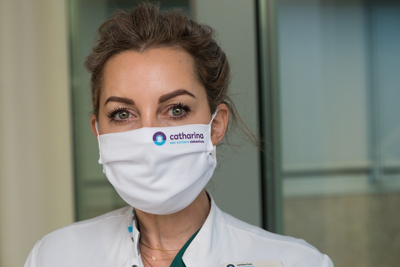 Vrouw met lichtbruin haar, mondmasker met Catharina-logo en witte jas, spreekt binnen over huidverzorging en maskné.