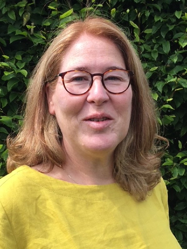 Vrouw met halflang lichtbruin haar en bril, draagt geel shirt, staat buiten voor groen struikgewas en kijkt licht glimlachend.