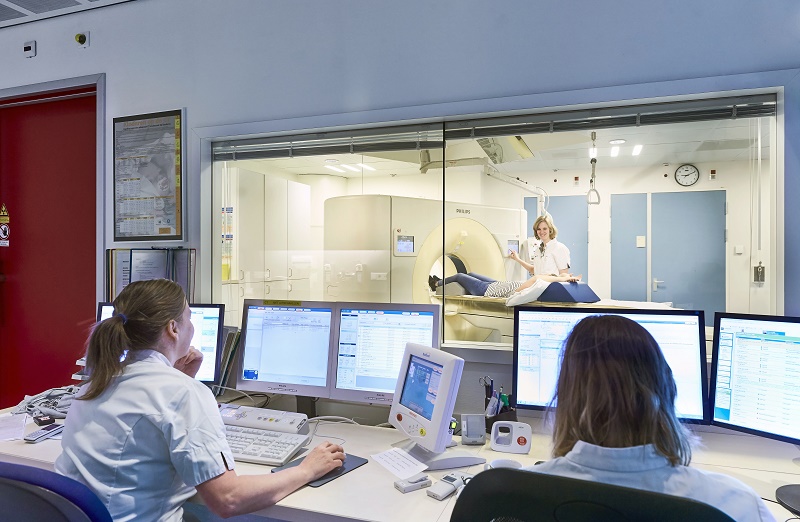 Twee medewerkers bedienen geavanceerde scantechniek in de controlekamer terwijl een andere medewerker een patiënt voorbereidt op een MRI-scan.