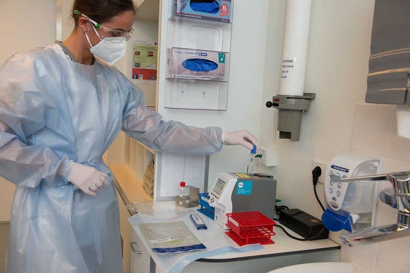 Persoon in beschermende kleding werkt met laboratoriumapparatuur in Catharina Ziekenhuis, buisjes en sneltest op werkbank.