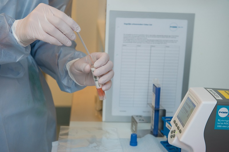 Persoon met handschoenen en labjas gebruikt een pipet bij sneltest; labapparaat en formulier op de achtergrond in Catharina Ziekenhuis.