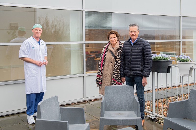 Drie volwassenen buiten bij een gebouw met grote ramen; één verpleegkundige apart, twee in winterkleding bij de reling met bloempotten.