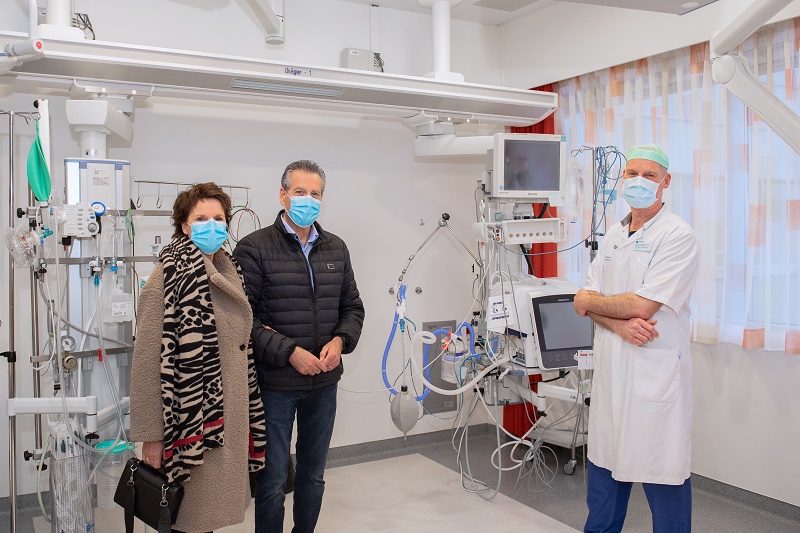 Twee bezoekers met jassen en maskers en een verpleegkundige in scrubs geven extra zorg aan een IC-patiënt in de ziekenhuisruimte.