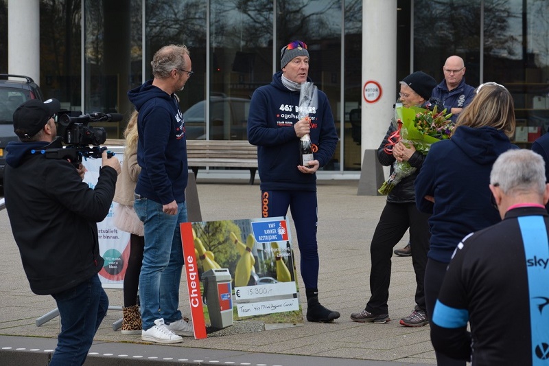 Groep mensen voor gebouw. Man in sportkleding spreekt en houdt fles vast, vrouw van Catharina Ziekenhuis met bos bloemen, grote cheque voor kankeronderzoek.