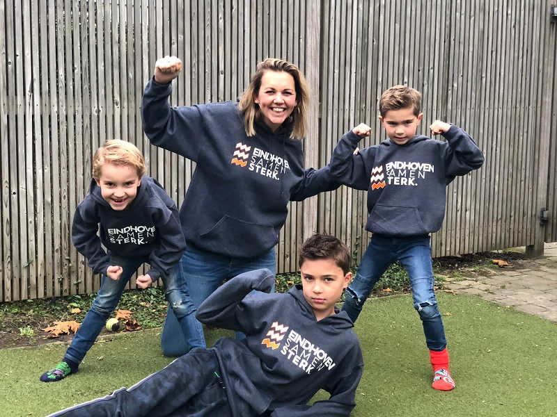 Vrouw en drie jongens in Eindhoven Samen Sterk truien poseren op kunstgras voor een houten schutting; één jongen ligt ervoor.