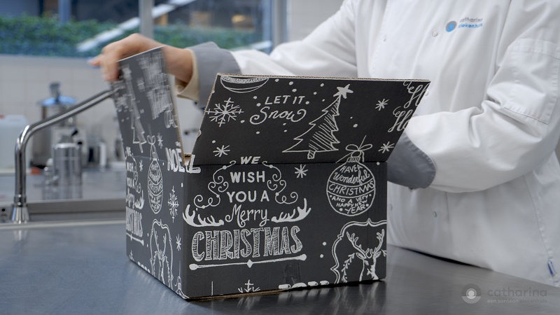 Een persoon in witte labjas opent een kerstdoos op een roestvrijstalen aanrecht. De doos heeft witte tekeningen en teksten.