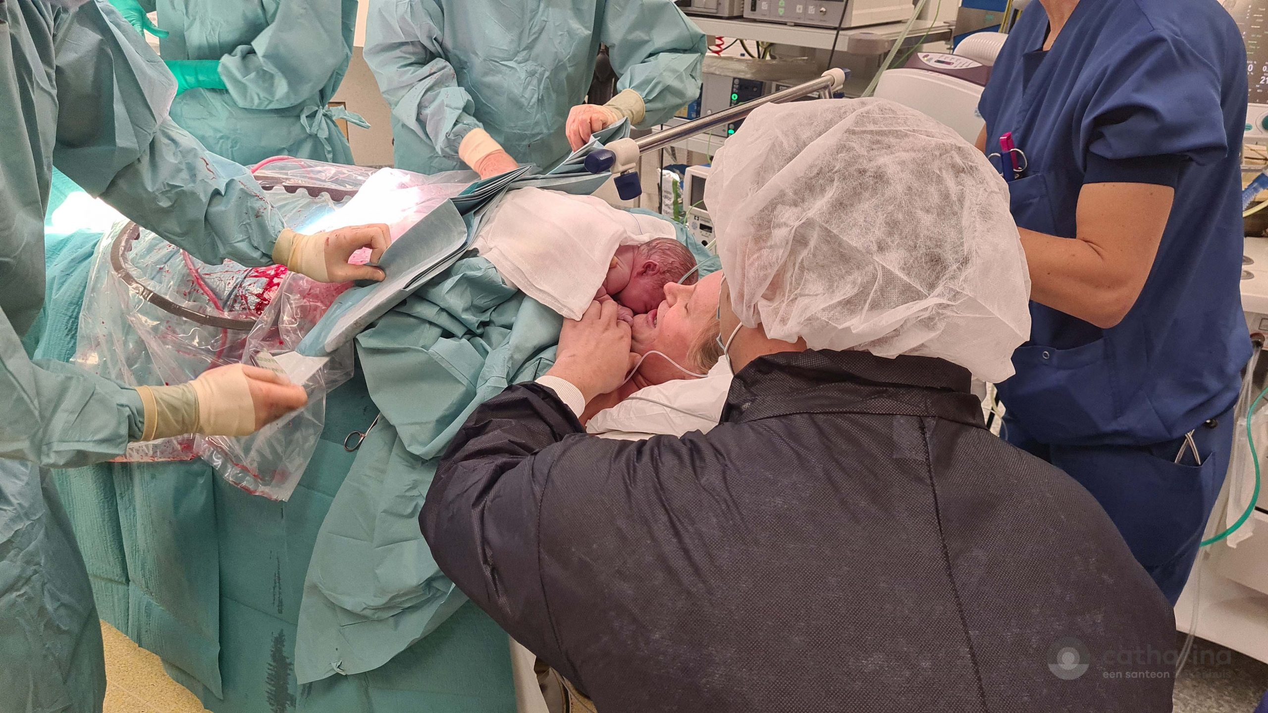 Een pasgeboren baby wordt bij Moeder Kelly gehouden tijdens een keizersnede, omringd door medisch personeel in operatiekamer.