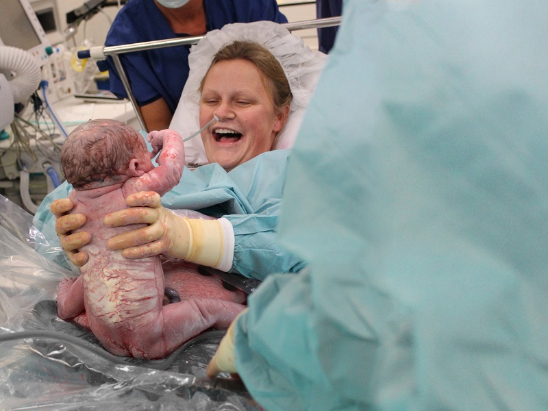 Een pasgeboren baby wordt vastgehouden door een medisch professional; moeder Kelly glimlacht op bed na de geboorte van Guus.
