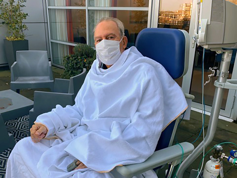 Persoon met wit deken en mondkapje zit buiten in een ligstoel naast IC-apparatuur. Tuinmeubilair en planten op de achtergrond.