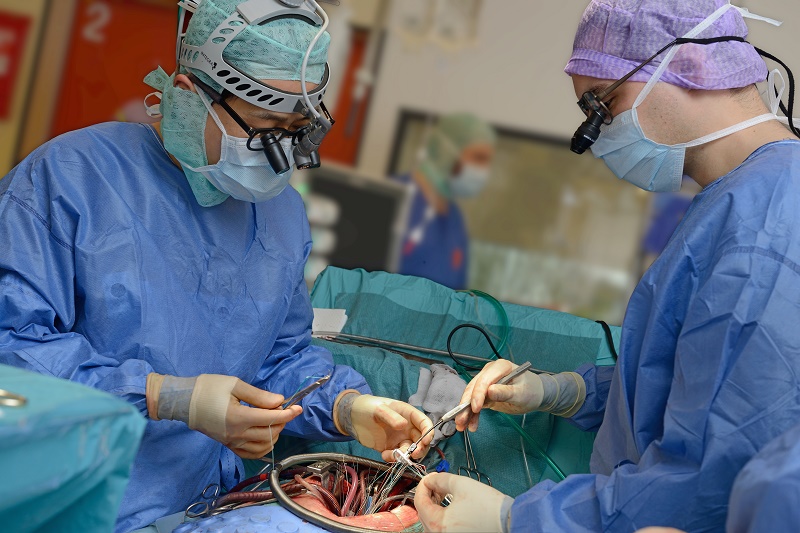 Twee chirurgen in blauw voeren een hartoperatie uit op een vrouw; instrumenten, slangen en een assistent op de achtergrond.