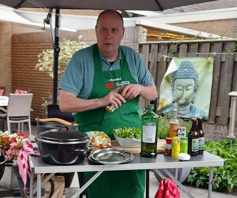 Ronald met groene schort bereidt eten aan buitentafel met ingrediënten; een Boeddha-kunstwerk hangt aan de schutting.