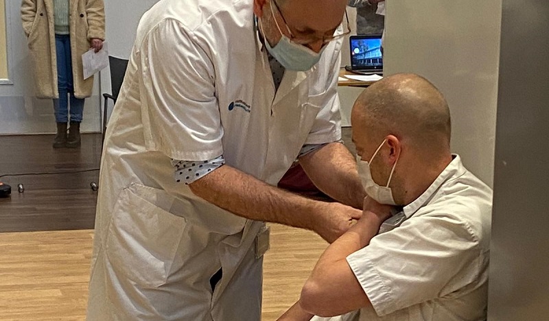 Een zorgmedewerker met mondkapje geeft een vaccinatie aan een zittend persoon in het Catharina Ziekenhuis; iemand staat bij de deur.