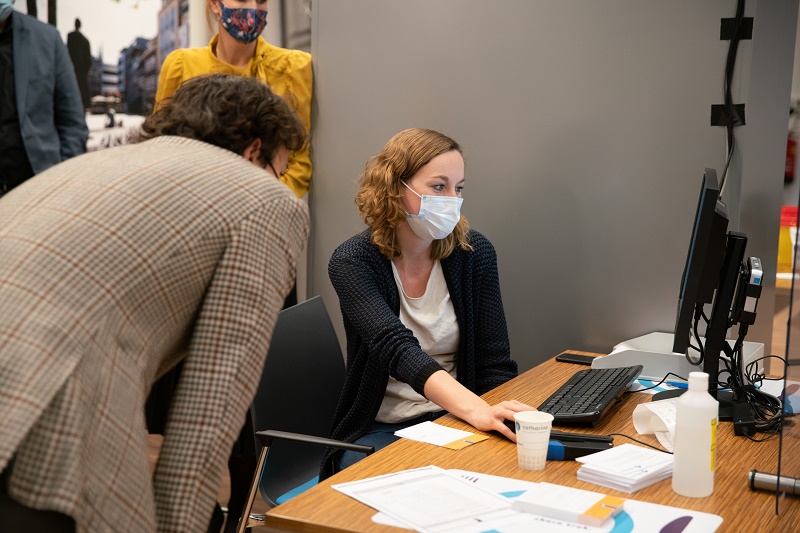 Vrouw met mondkapje werkt achter computer en wijst op scherm. Man leunt mee. Papieren op bureau, mensen staan op achtergrond.
