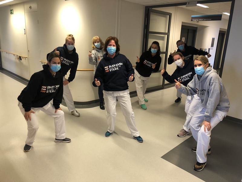 Zeven personen met mondmaskers, donkere hoodies en witte broeken poseren op afstand in een ziekenhuisgang in Eindhoven.