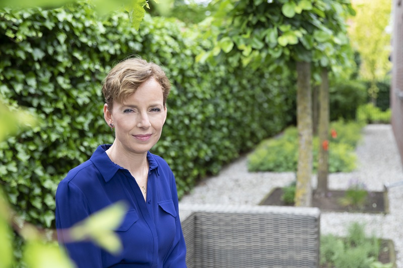Vrouw met kort lichtbruin haar en blauwe blouse staat in een tuin met groene hagen, grindpaden en kleine bomen.