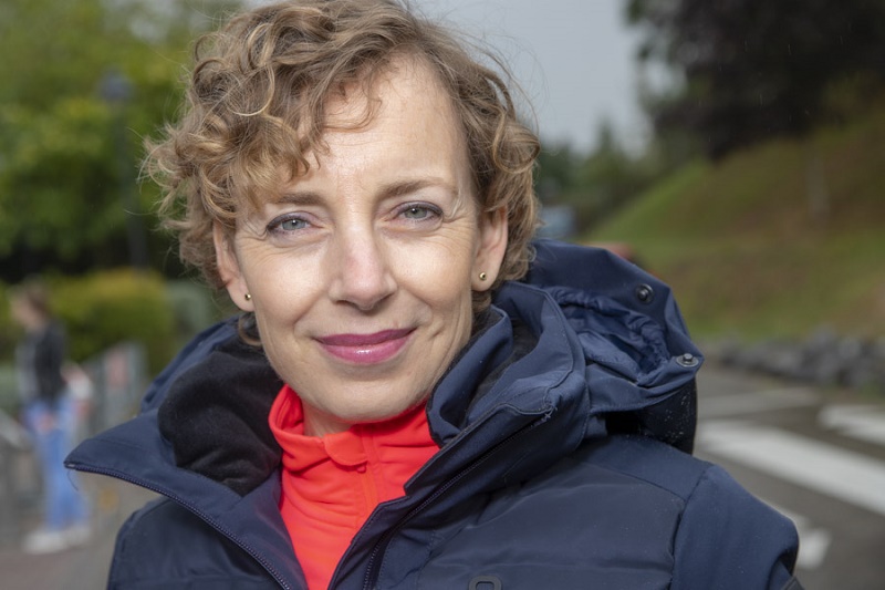 Vrouw met kort krullend haar en blauwe jas staat buiten, kijkt naar de camera. Op de achtergrond groen en een zebrapad.