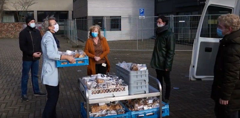 Vijf mensen met mondkapjes, mogelijk zorgpersoneel van Catharina Ziekenhuis, verdelen verpakt eten buiten bij een gebouw en busje.