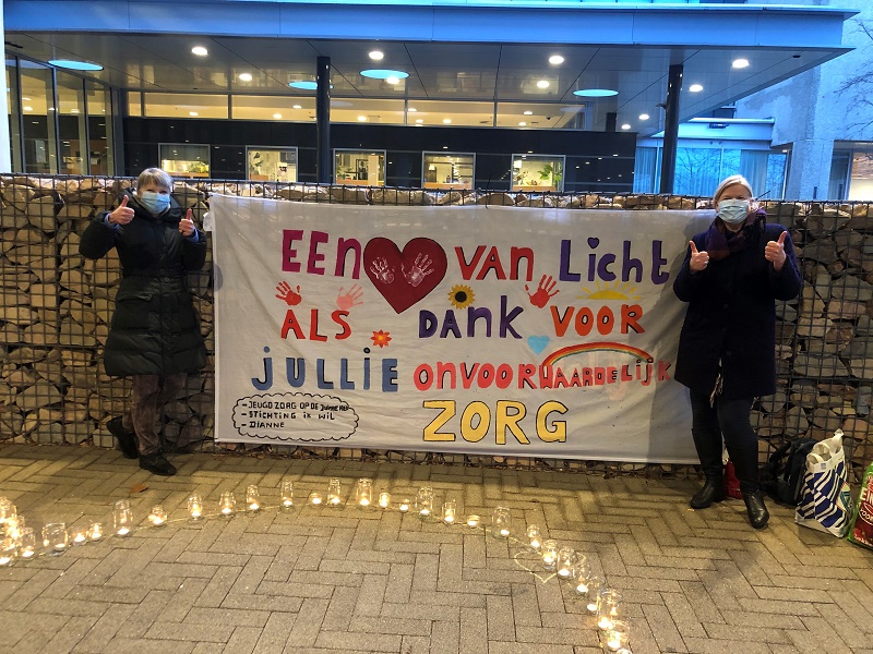Twee mensen met mondkapjes staan bij een groot spandoek voor zorgpersoneel, geven duimen op, kaarsen branden voor hen.