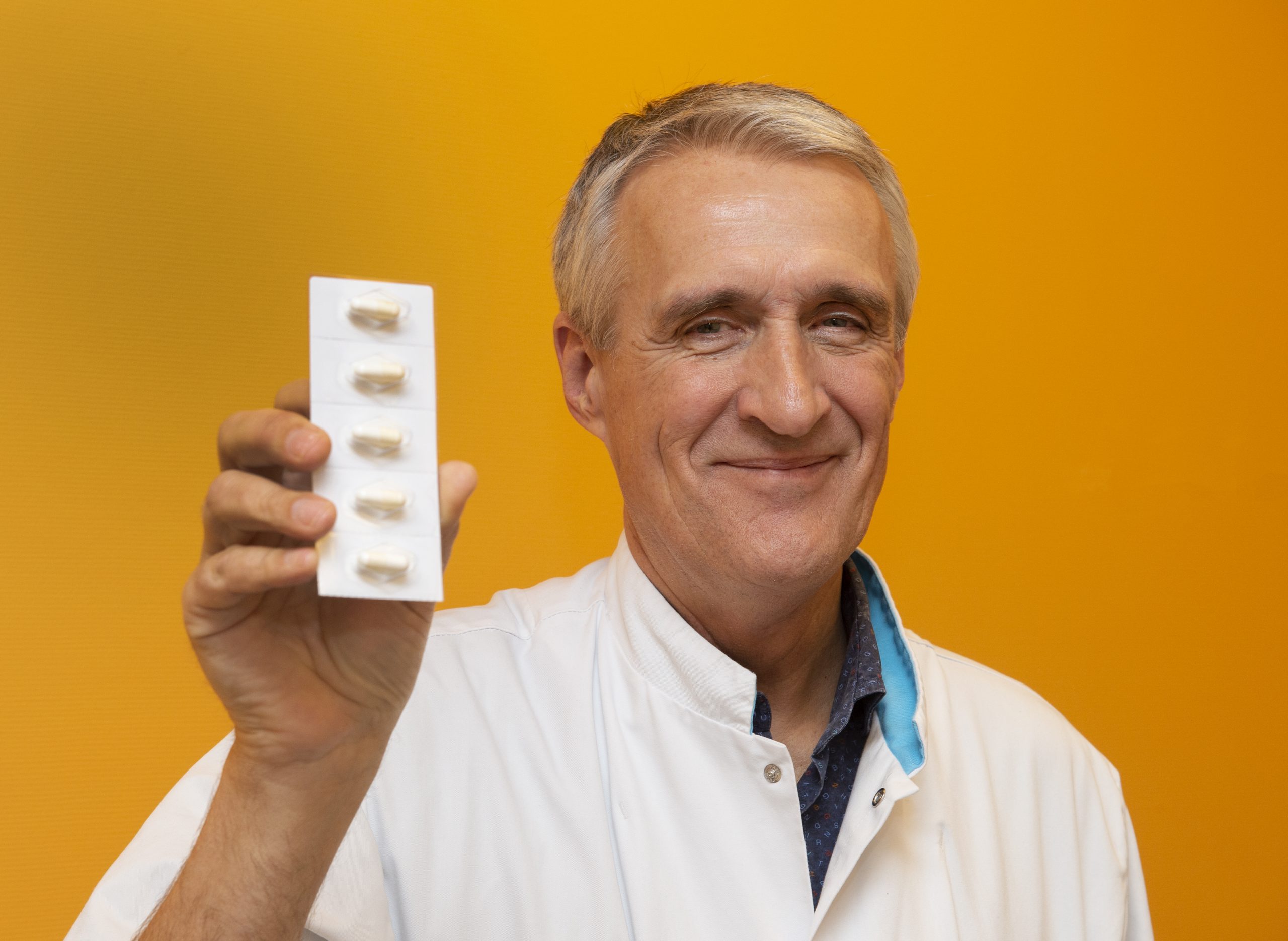 Man in witte labjas toont een blister met zes capsules voor MS-behandeling thuis, tegen een oranje-gele achtergrond.