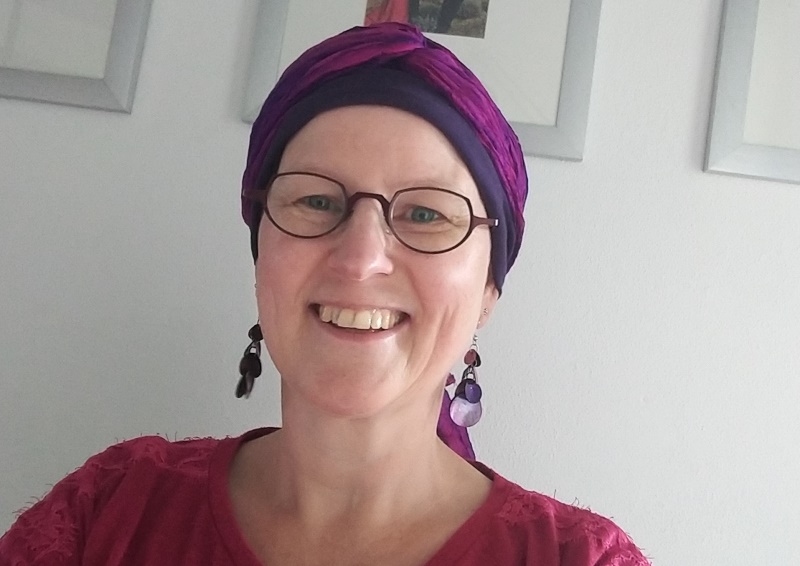 Persoon met bril, paarse en roze hoofddoek en oorbellen glimlacht naar de camera. Roze top, witte muur met fotolijsten.