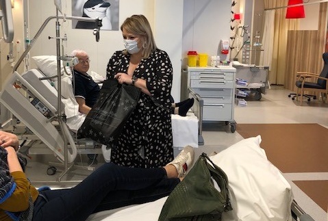 Vrouw met mondkapje bij ziekenhuisbed met goodiebag, man in ander bed. Medische apparatuur zichtbaar op Wereldkankerdag.