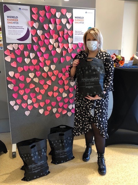 Vrouw met mondkapje voor bord met roze en rode harten. Zij houdt een zwarte goodiebag vast; twee liggen op de grond. Posters over Wereldkankerdag en dagbehandeling zichtbaar.
