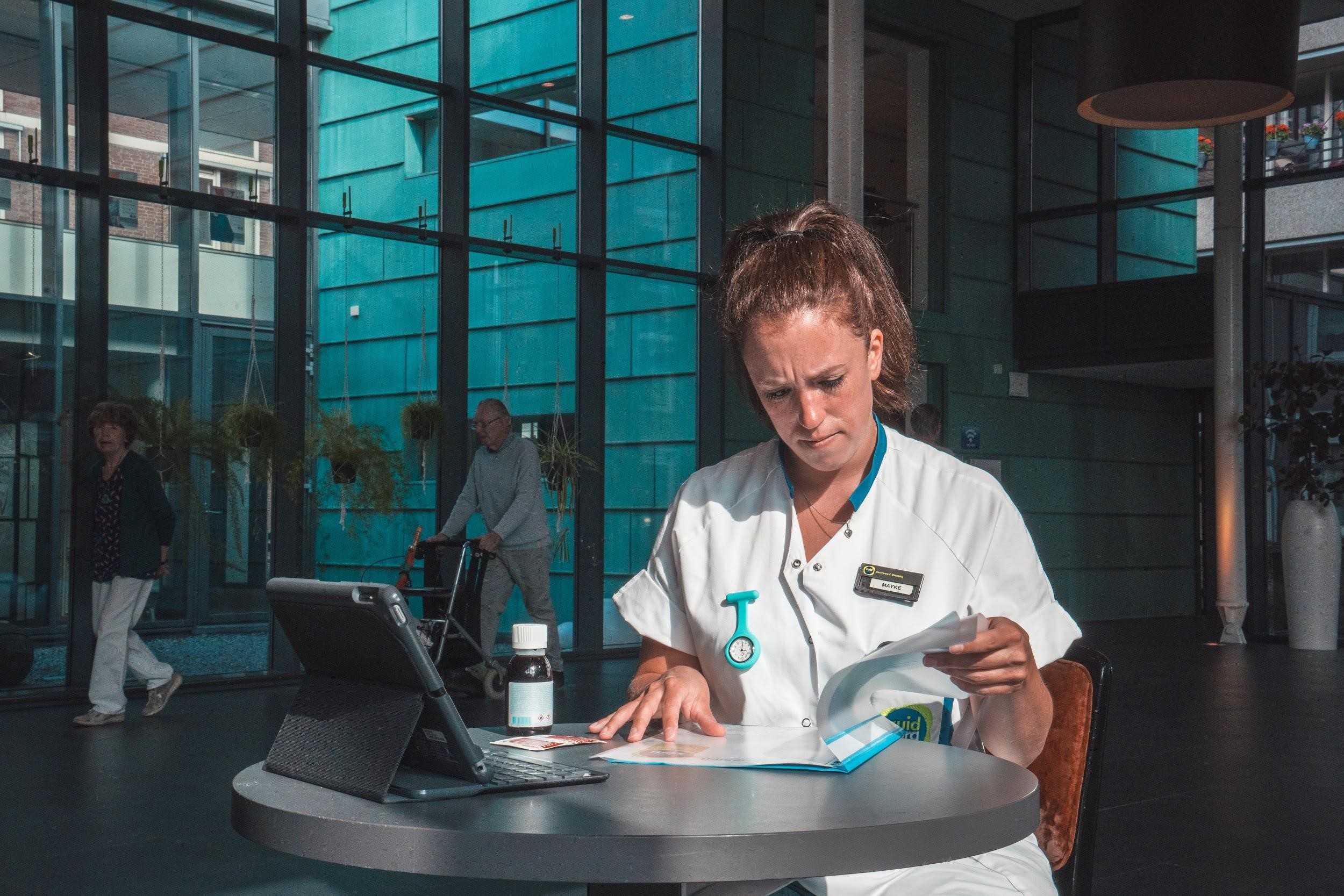 Vrouw in witte medische kleding werkt aan tafel met papieren en tablet in modern ziekenhuis, anderen zichtbaar achter glas.
