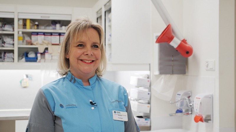 Vrouw in lichtblauw medisch uniform met naamkaartje in moderne kliniekkamer van Catharina Ziekenhuis, met medische spullen.