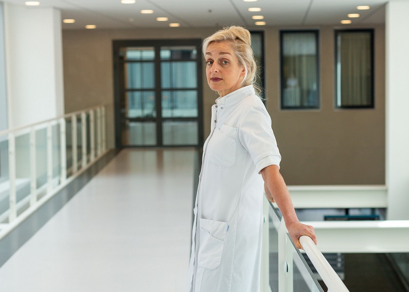 Vrouw in witte labjas kijkt over haar schouder in een moderne, helder verlichte gang en leunt met één hand op de reling.