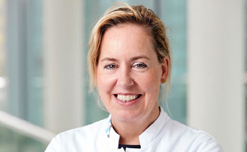 Verloskundige Simone. Ze staat binnen, met grote ramen op de achtergrond.