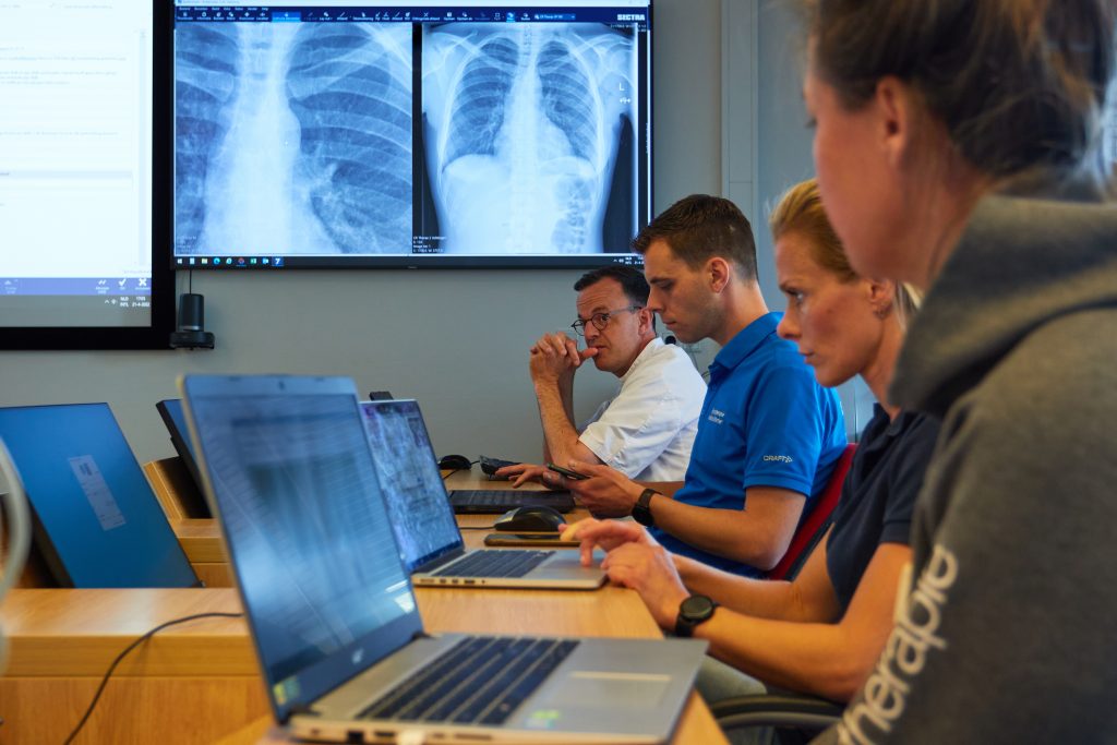 Vier mensen werken aan laptops; op de achtergrond toont een monitor twee thoraxfoto's. Eén persoon draagt een witte jas.