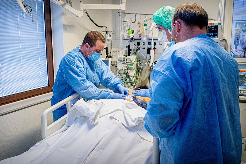 Drie zorgverleners in beschermende kleding helpen een patiënt in een ziekenhuisbed, omringd door medische apparatuur.