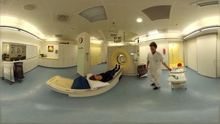 Een persoon ligt op een CT-scantafel in een heldere ziekenhuiskamer; een zorgverlener bereidt apparatuur voor.