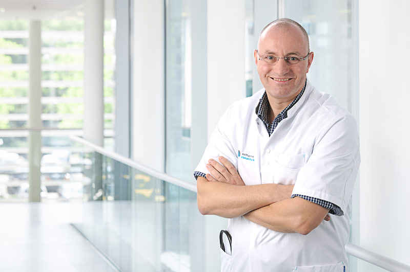 Man met bril en witte jas, gespecialiseerd in nieuwe behandelingen en medicijnen, staat lachend in lichte, moderne gang.