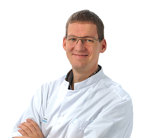 Een man met bril en witte labjas staat tegen een witte achtergrond, gekruiste armen, en kijkt glimlachend in de camera.