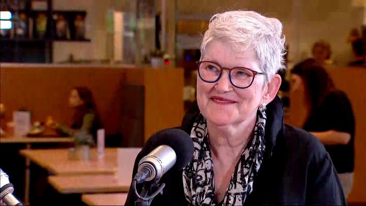 Gerda Cobben zit glimlachend achter een microfoon, in zwarte outfit met sjaal. Achter haar zit een café vol mensen aan tafels.