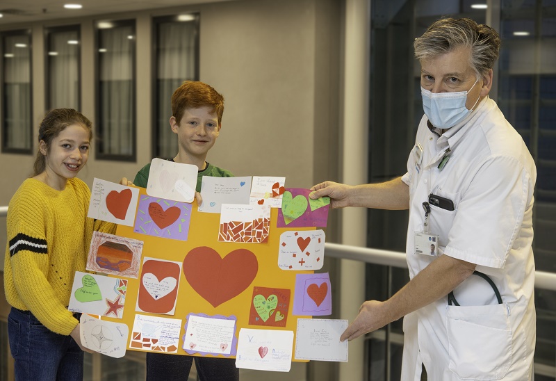 Twee kinderen en een zorgmedewerker met mondkapje houden in het ziekenhuis een gele poster van basisschool De Regenboog vast.