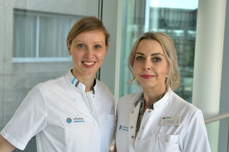 Twee vrouwen in witte medische uniformen staan bij een groot raam in een modern gebouw. Ze hebben licht haar en dragen badges.