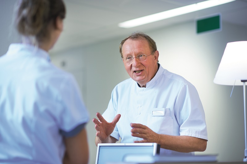 Een mannelijke arts in witte jas bespreekt met handgebaren de onderzoeksresultaten met een vrouwelijke collega in een lichte kliniek.