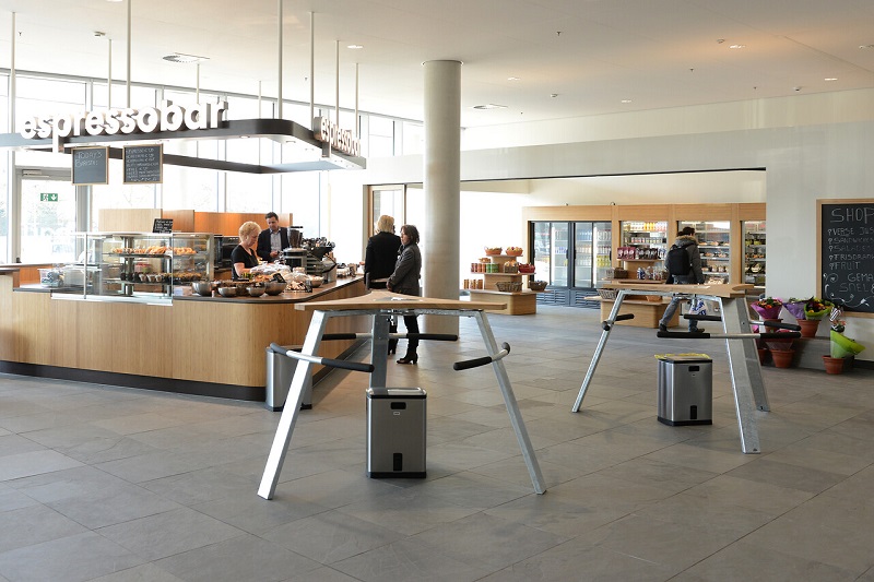 Modern, ruim bezoekersrestaurant met espressobar. Mensen bestellen drankjes. Houten statafels, schappen met boodschappen en planten.