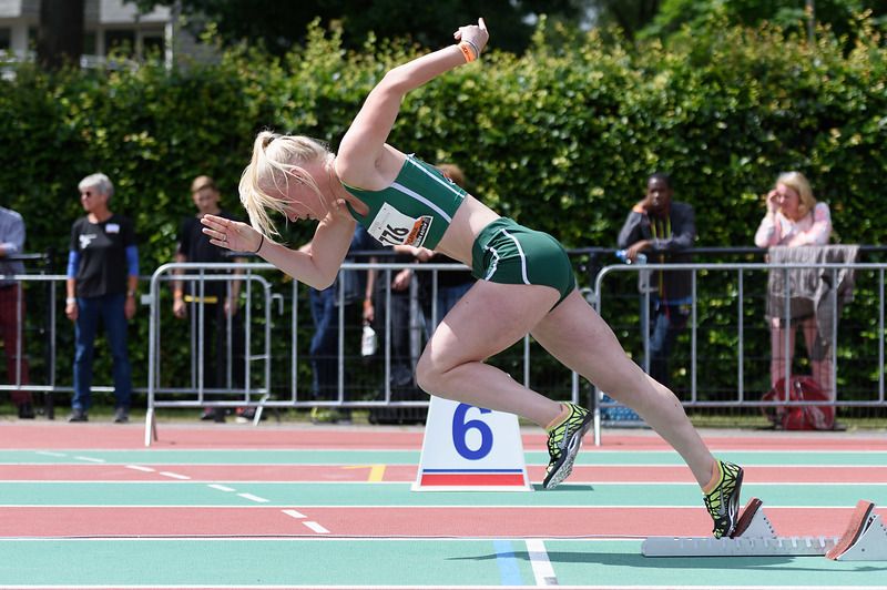 Vrouwelijke sprintster in groen-wit start race vanuit baan 6 op buitenbaan; toeschouwers achter metalen hek.