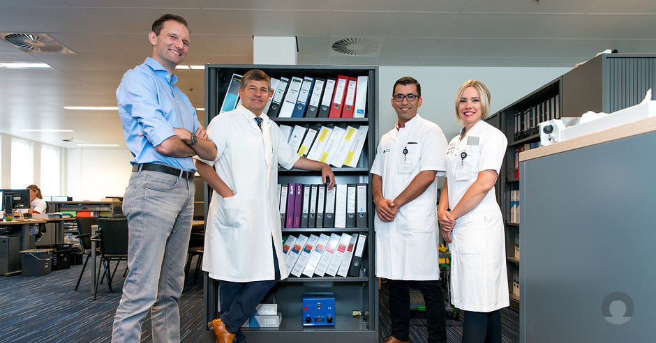 Vier mensen in een kantoor bij ordners; drie in witte jassen bespreken onderzoek, één in blauw staat ernaast. Bureaus op achtergrond.
