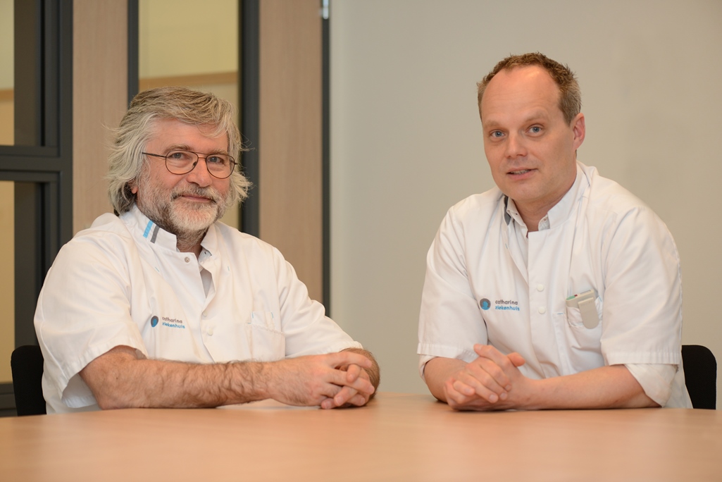 Twee mannen in witte jassen zitten aan een tafel in een klinische ruimte met neutrale muren, kijkend naar de camera.