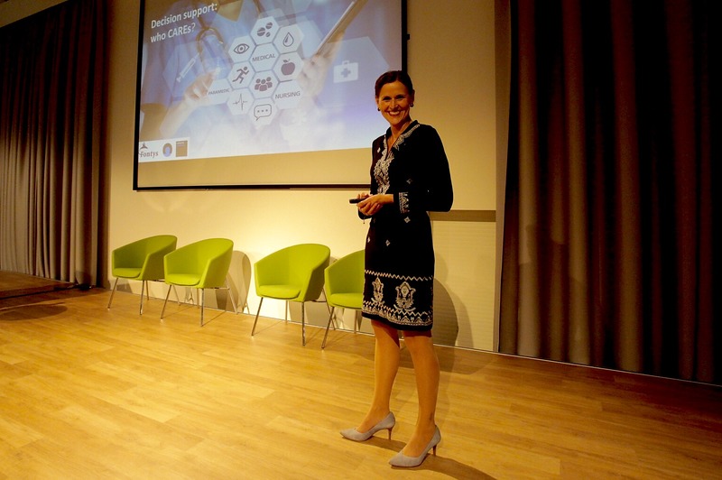 Vrouw in zwart-witte jurk staat op podium met afstandsbediening, drie groene stoelen en presentatie over slimme zorgtechnologie.