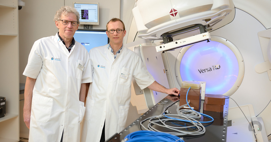 Twee personen in witte labjassen staan naast het bestralingstoestel Versa HD in een kliniek, omringd door kabels en monitoren.