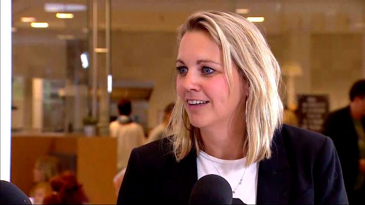 Vrouw met blond halflang haar, zwart colbert en witte blouse spreekt binnen over darmkanker. Wazige achtergrond met mensen.
