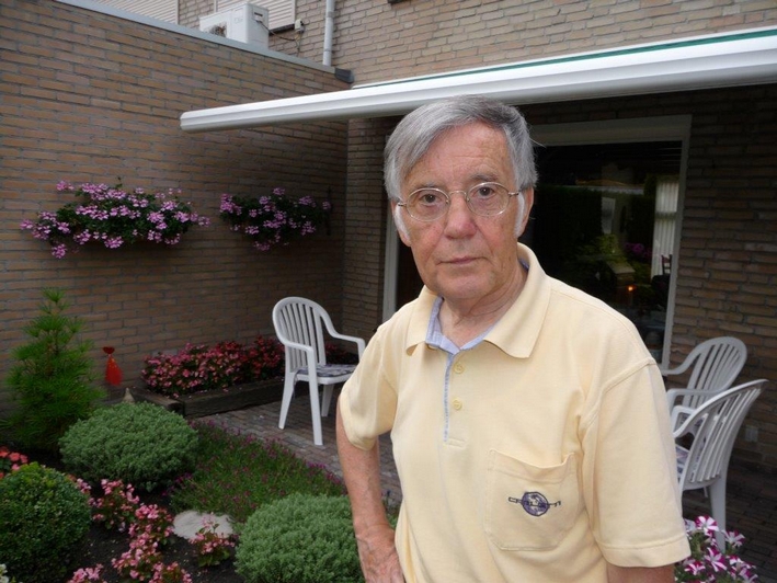 Oudere man met grijs haar en bril staat in bloementuin. Gele polo, ontspannen na borstoperatie. Witte stoelen en huis achter hem.