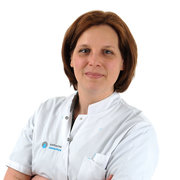 Persoon met kort bruin haar en witte doktersjas staat met gekruiste armen en glimlacht licht tegen een witte achtergrond.